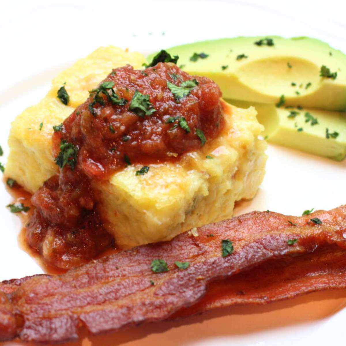 A plated piece of delicious green chili egg casserole with two pieces of crispy bacon and sliced avocado on the side. The breakfast casserole is piled high with homemade salsa and chopped cilantro as its being served.