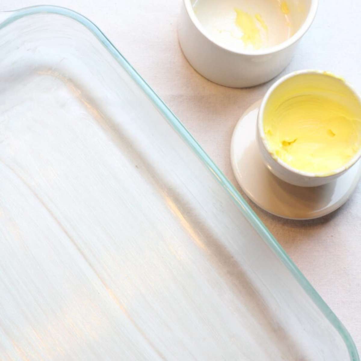 A large clear baking dish that has been lightly greased with homemade butter. A white butter bell sits next to the baking dish with bright yellow butter inside. 