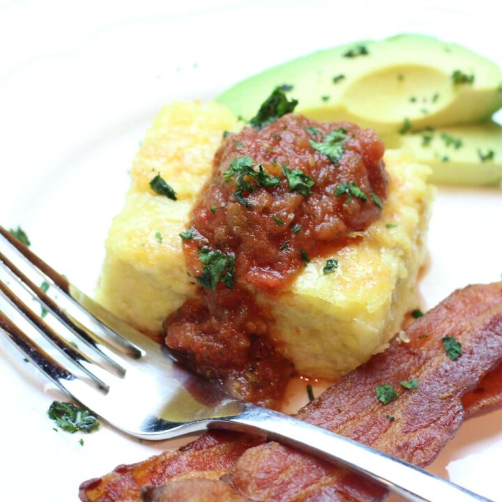 A white plate piled high with Green Chile Breakfast Casserole. Topped with homemade fresh salsa and minced cilantro. Two strips of crispy bacon and sliced avocado are resting on the same place ready for breakfast.