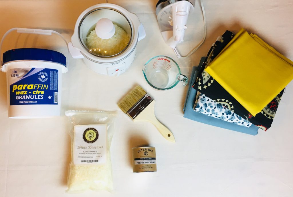 A painters drop cloth sitting on a table with all the supplies to wax a few yards of canvas fabric material. Paraffin wax, beeswax pellets, paintbrushes, a miniature white crockpot and an iron neatly placed on the table.