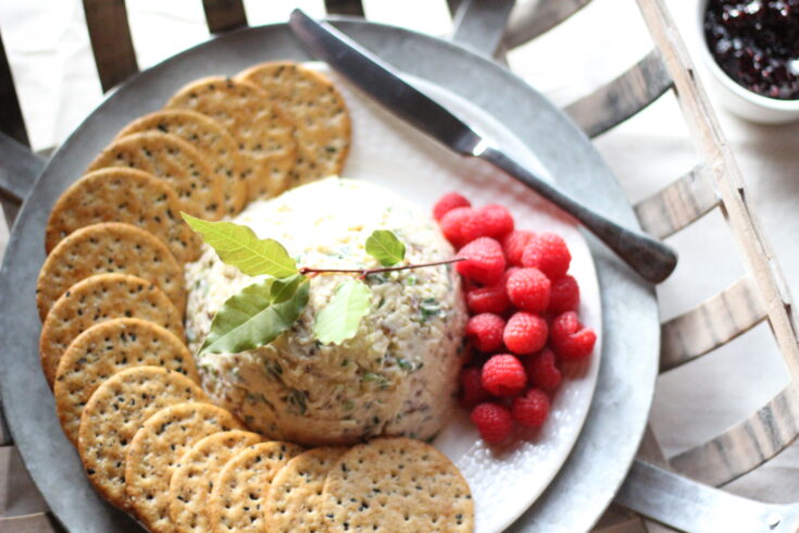 A delicious white savory cheese spread appetizer placed neatly in a half dome shape on a white plate. Next to the cheese spread sits a small pile of red raspberries and some multigrain crackers. Placed on top of the cheese spread is a green bay leaf spring. A silver knife rests on the plate nearby.