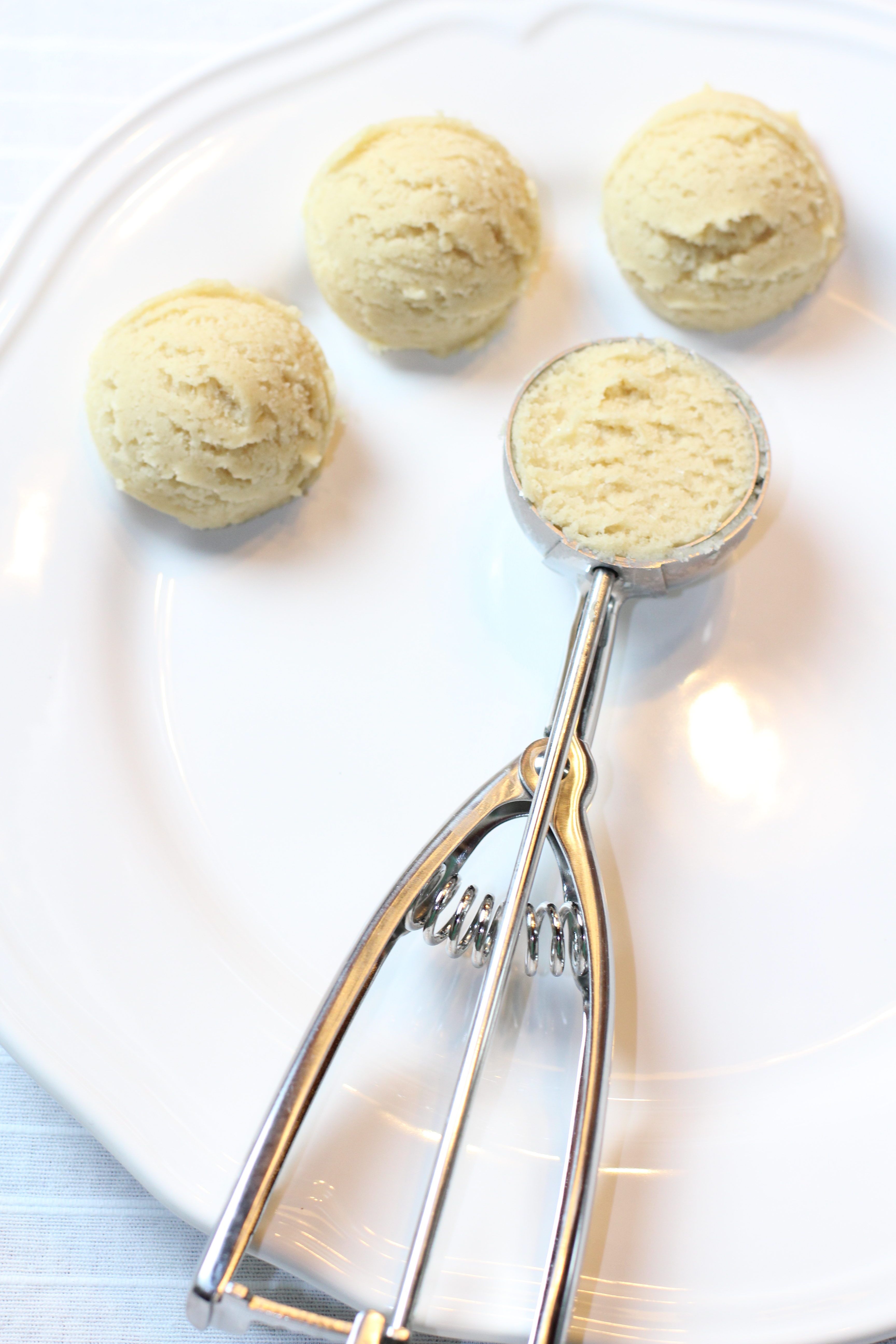 A white plate with a silver ice cream scoop used to scoop sugar shortbread dough from the stand mixing bowl.