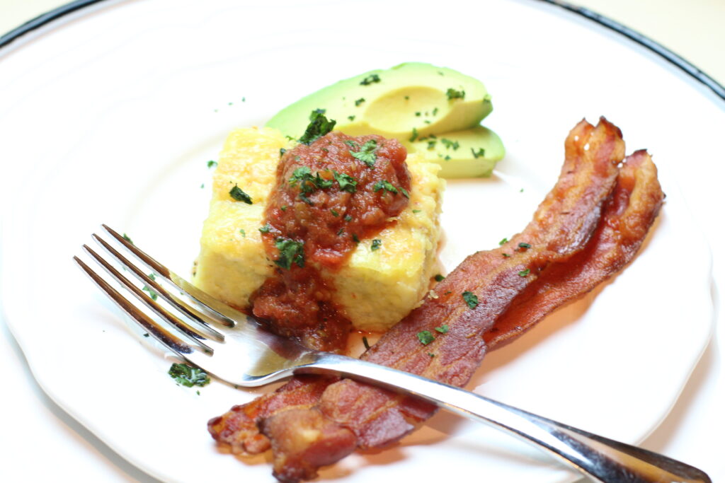 Green Chile egg casserole on white serving plate topped with homemade fresh salsa and minced cilantro. Two strips of crispy bacon and sliced green avocado are on the plate beside the casserole.