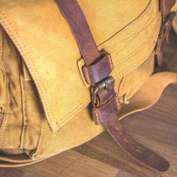 a mustard yellow waxed canvas bag with dark brown leather straps is sitting on a wooden table. The picture shows only the left corner front of the bag.