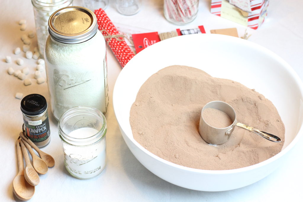 A freshly mixed batch of hot cocoa mix sitting is a white bowl. A silver measuring cups rests inside the bowl to scoop the mix into mason jars for gifts. 