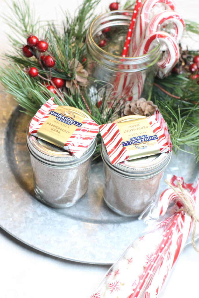 A silver plate with two mason jars full of homemade hot cocoa mix. Some red and white striped peppermint white chocolates sitting on the tops of the jars. Christmas greenery is sitting nearby.
