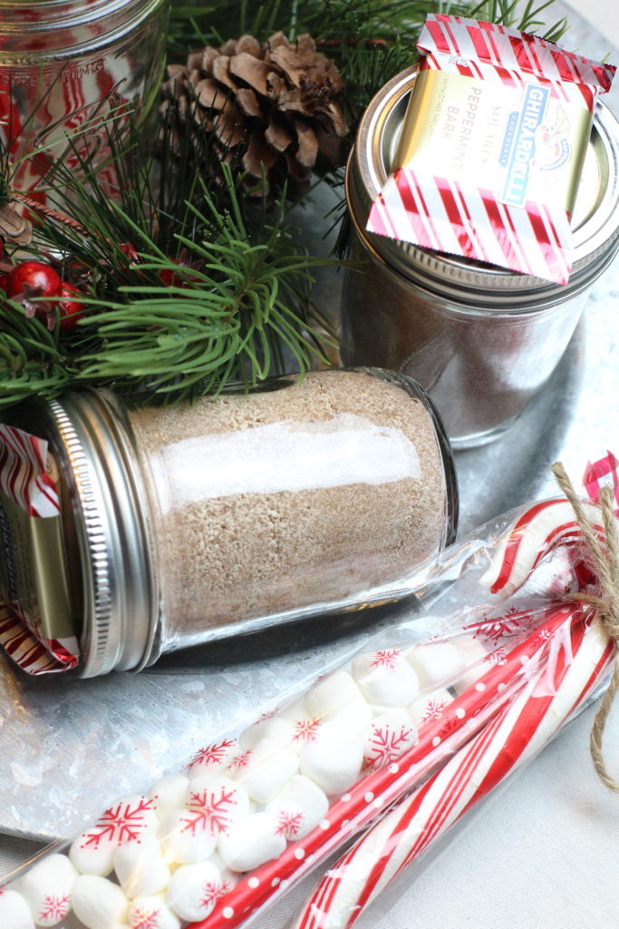 A jar of homemade hot chocolate mix is laying on it's side ontop of a silver platter. A small clear bag of mini marshmallows, a red paper straw and a candy cane is tied with twine to secure it to the jar.