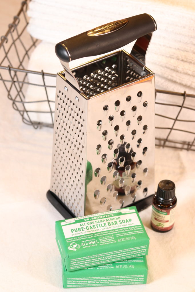 A silver metal box grater sitting in front of a basket of neatly folded white wash rags.