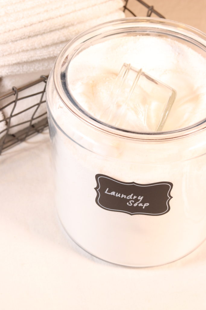 A clear glass container filled with white homemade laundry detergent powder. A metal basket of white washcloths is folded nearby.