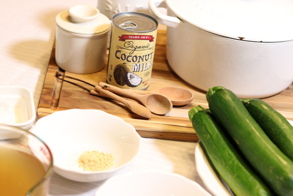 A table full of ingredients to make a silky zucchini soup recipe. All the ingredients are sitting on a wooden cutting board with a large white cast iron stock pot in the background.