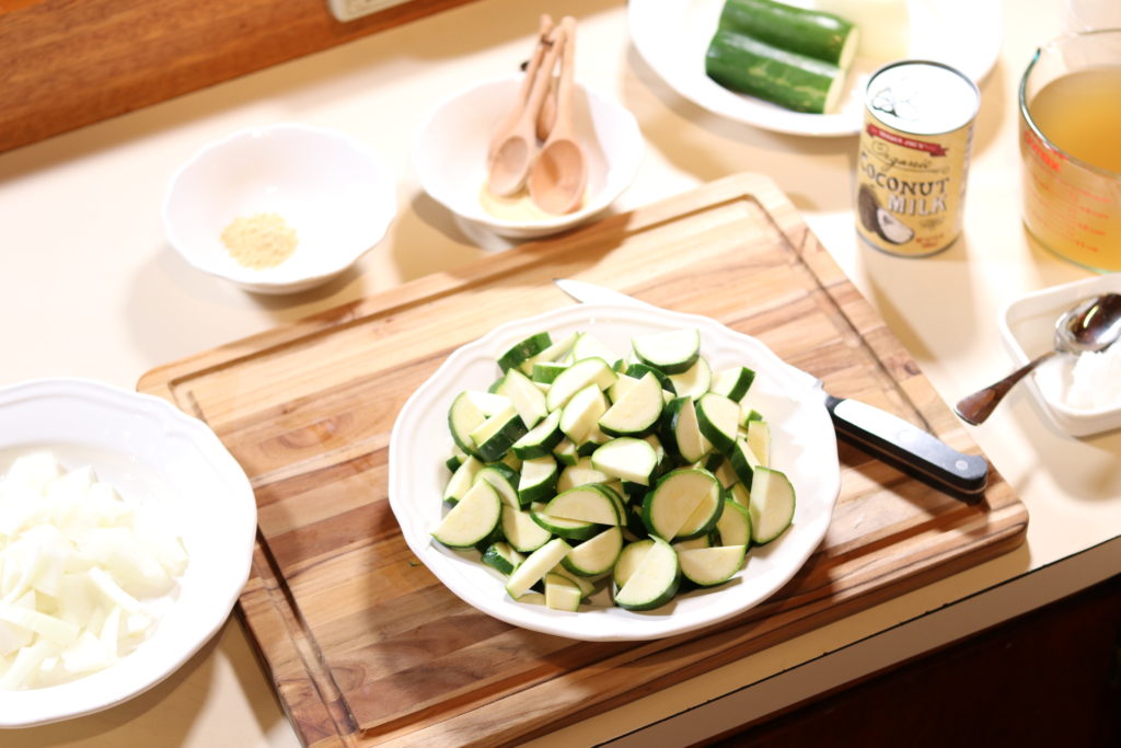 A freshly cut bowl of zucchini prepared for zucchini soup recipe. A can of coconut milk, a chopped onion and chicken stock rests nearby.