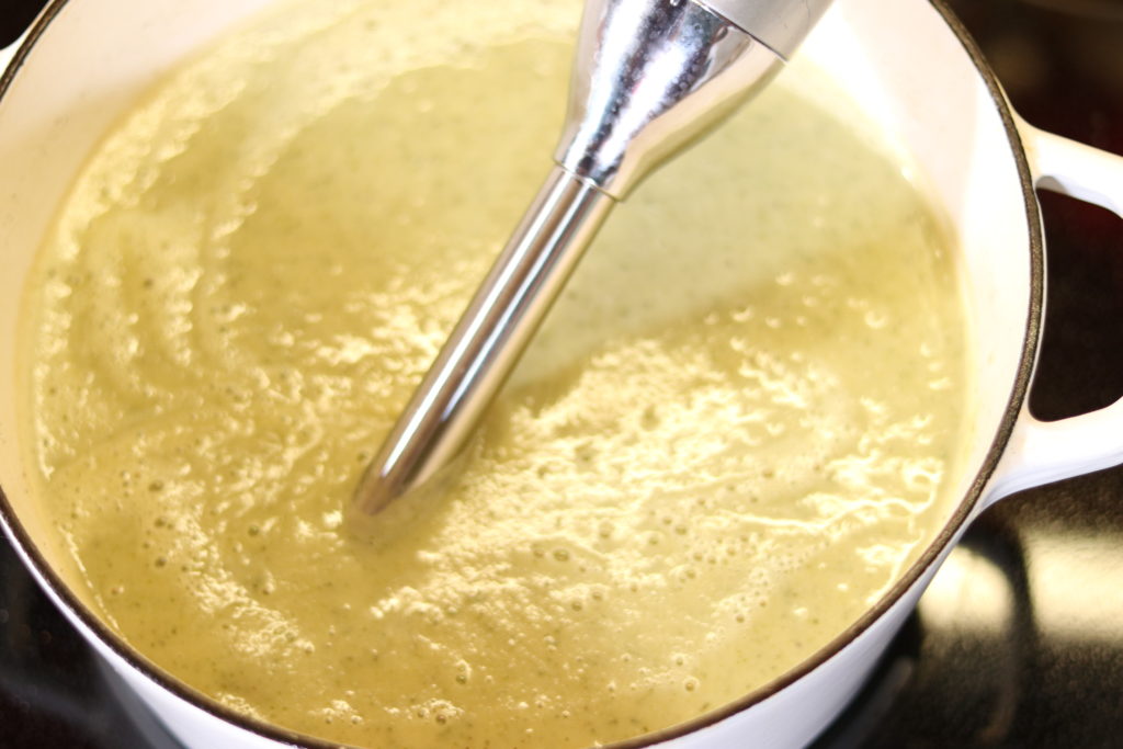 A silver immersion blender wand is sitting in a white stock pot of silky zucchini soup after blending. The soup is light green in color with small bubbles at the surface from heating.