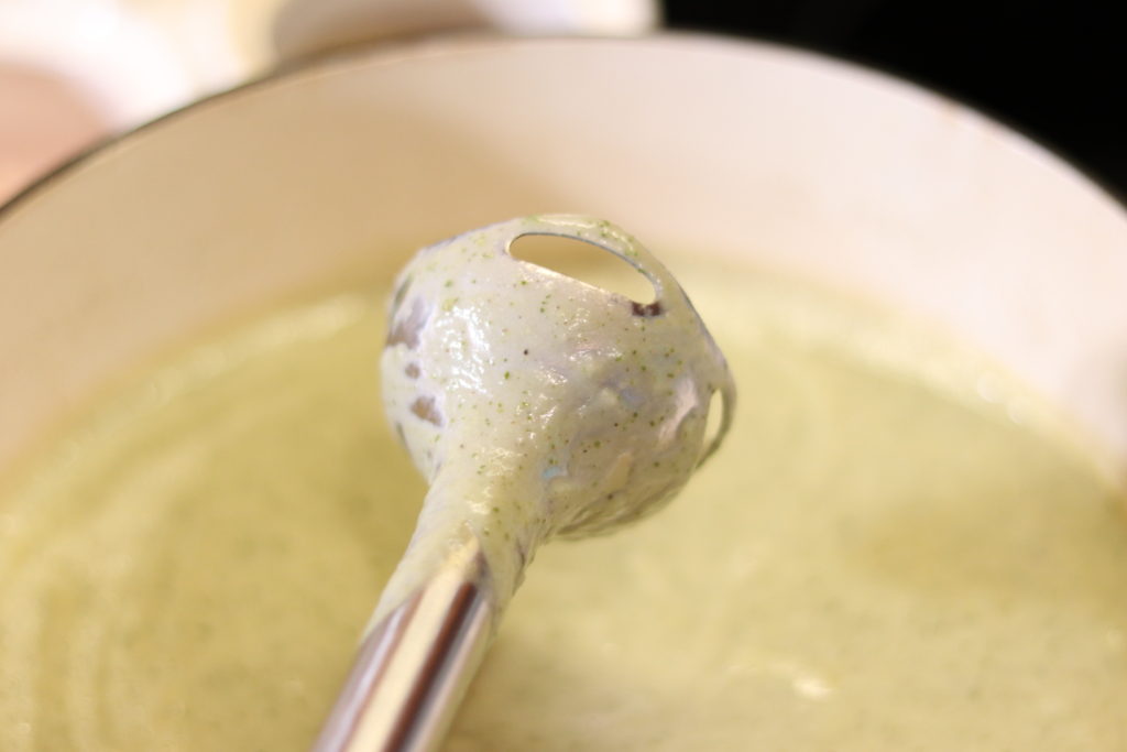 An immersion blender wand lifted from a pot of hot zucchini soup showing the texture and color after the soup has been blended.