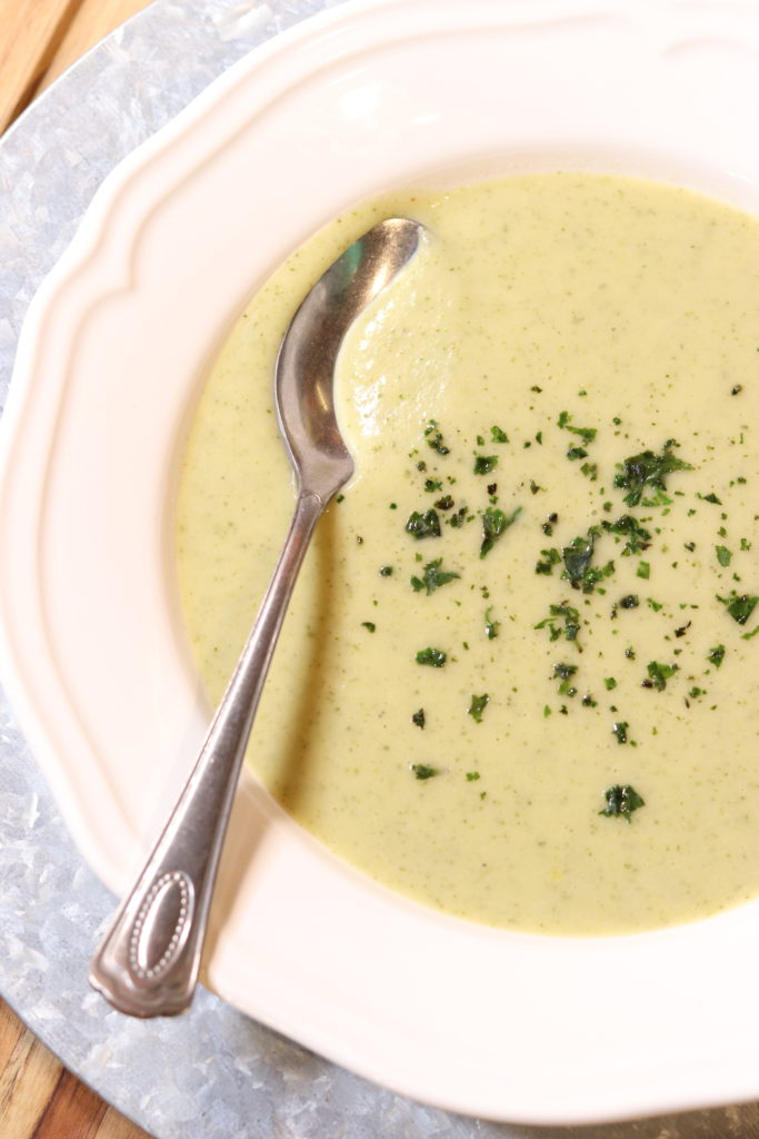 A bowl of silky zucchini soup served in a crisp white bowl. The soup is topped with minced chives. A silver spoon rests in the soup, ready for the first bite.