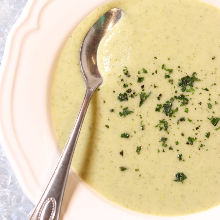 A white bowl filled with a light green creamy silky zucchini soup. A silver spoon rests in the soup and is topped with dried mince chives.