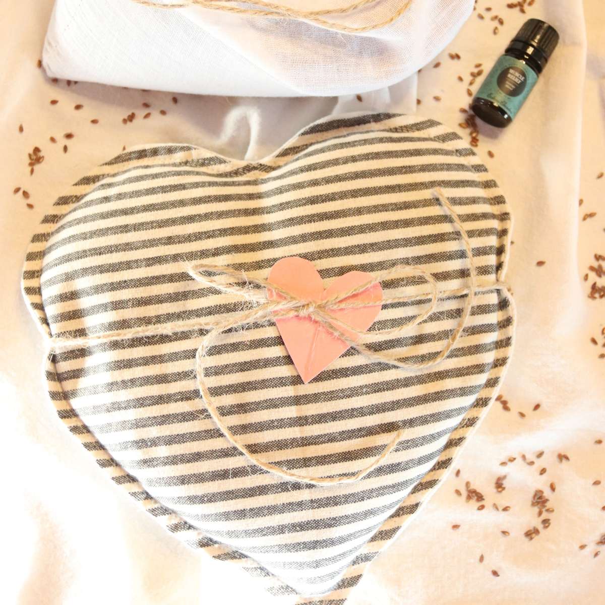 A heart shaped black and white reusable heating pad made with natural flax seeds and essential oils sitting on a white table cloth. The heart shaped heating pad is tied up with a piece of twine and flax seeds are scattered around nearby along with a bottle of essential oil.