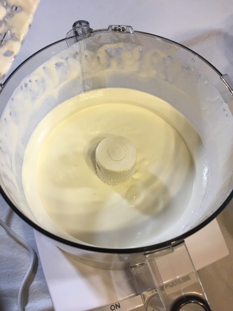 Heavy whipping cream is added to a large clear bowl of a food processor to begin making food processor butter.