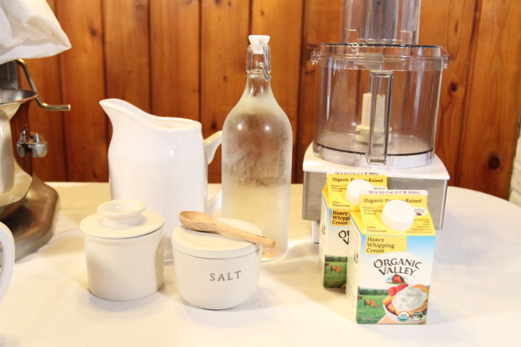 Food processor butter ingredients set on a table next to a white food processor. Two small containers of heavy whipping cream, salt and cold water are ready for making homemade butter.