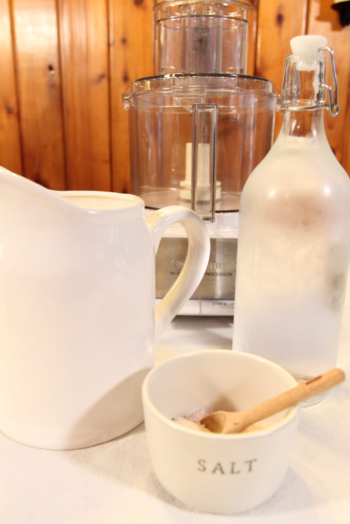 A large white food processor, a salt pot full of pink sea salt and a white pitcher full of chilled water rests on a table ready to make food processor butter.