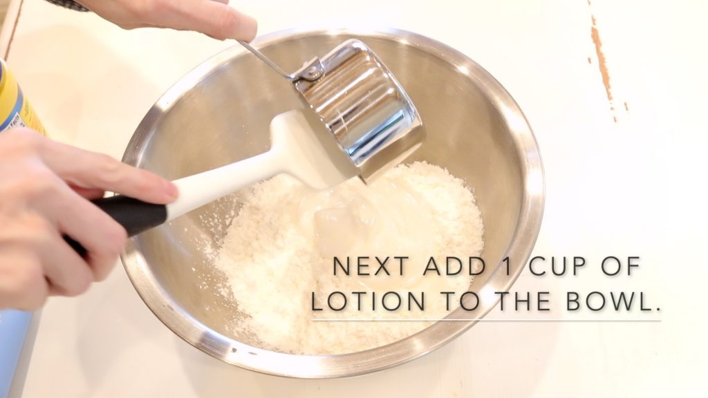 Adding hand lotion to the cloud dough recipe. A woman is holding the metal measuring cup and using a spatula to scrape out the cup into the bowl below.
