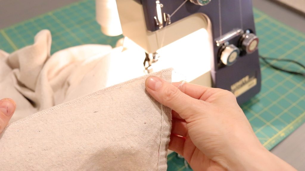 A woman holding an edge of canvas fabric that has just been sewn with a serger using white thread. The serger is sitting on the table in the background.