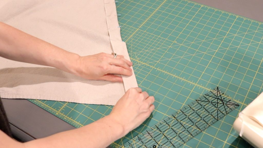Two hands finger pressing and pinning down the raw edge of a piece of fabric. The fabric is sitting on a green pressing mat with a clear ruler nearby. 