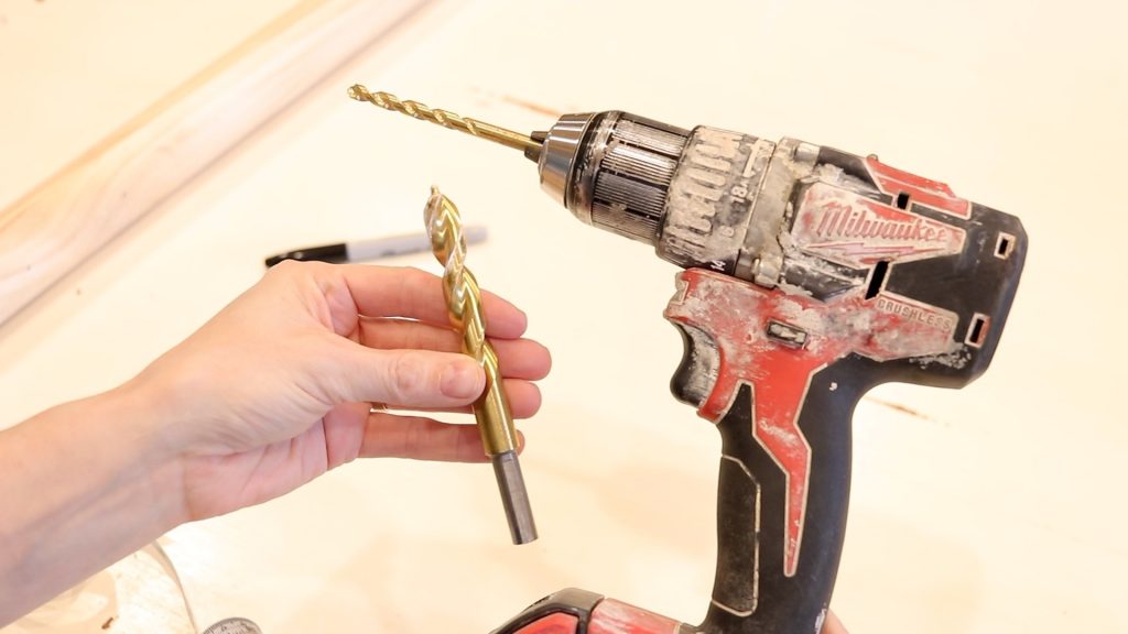 A hand is holding up a gold drill bit white the other hand holds the red and black drill. 