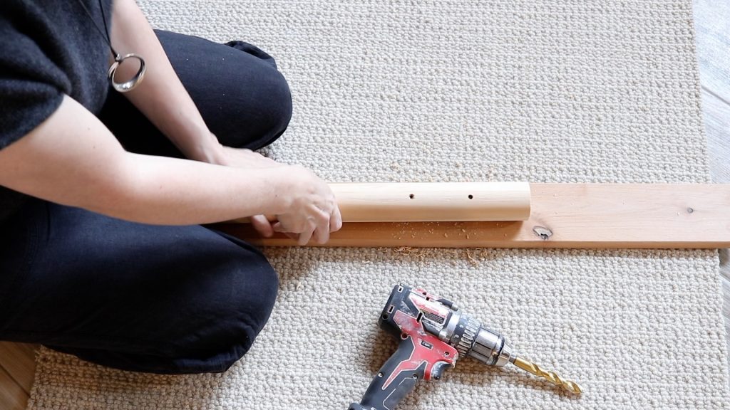 A woman wearing black pants and a black shirt is straddling a dowel rod preparing to drill the holes. There is a piece of wood under the dowel rod so the drill bit doesn't hit the floor while drilling. A red and black drill is sitting nearby. 