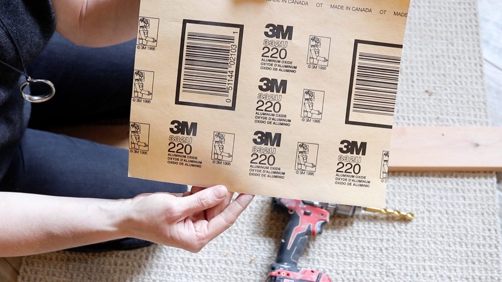 A woman is holding up a piece of 220 grit sand paper showing the back of the paper. A red and black drill is sitting on the floor next to her. 