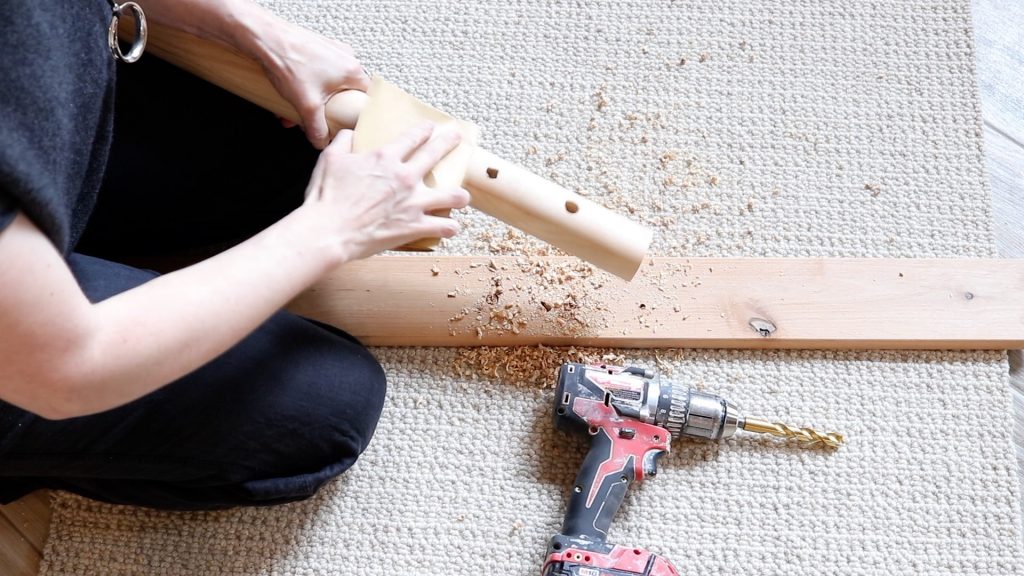 A woman wearing black is demonstrating how to sand the dowel rod after drilling. She is sitting on cream carpet with her drill nearby. 