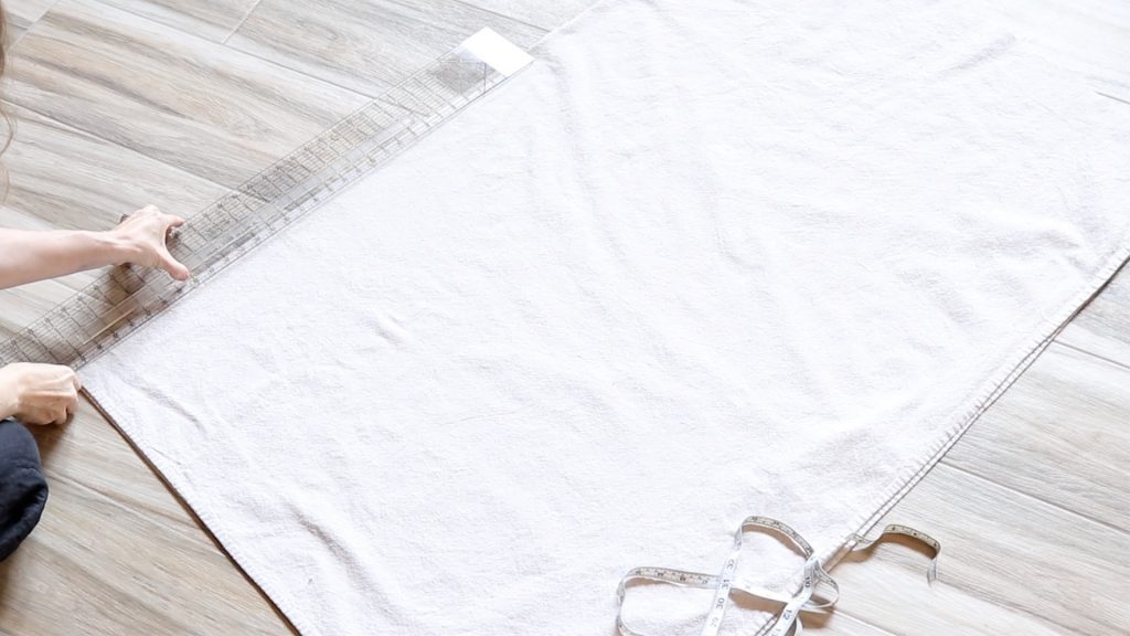 A large piece of canvas fabric laid out on the floor in preparation for cutting. The woman is holding a clear ruler and a white tape measure sits nearby. 