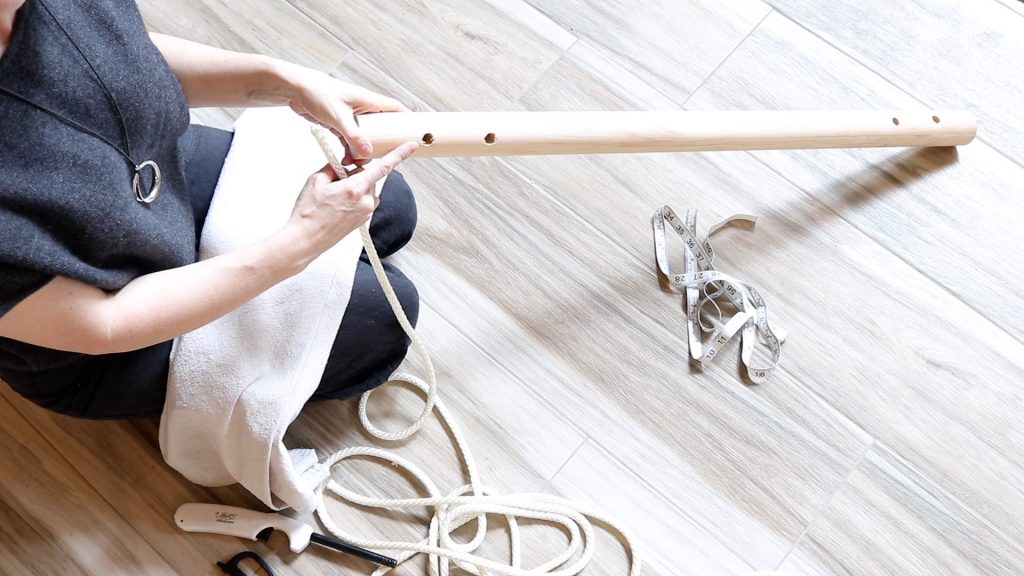 A woman sitting on the floor wearing black is holding a piece of white rope to thread through the hole on the wooden dowel rod. She is pointing to the hole the rope needs to pass through first.