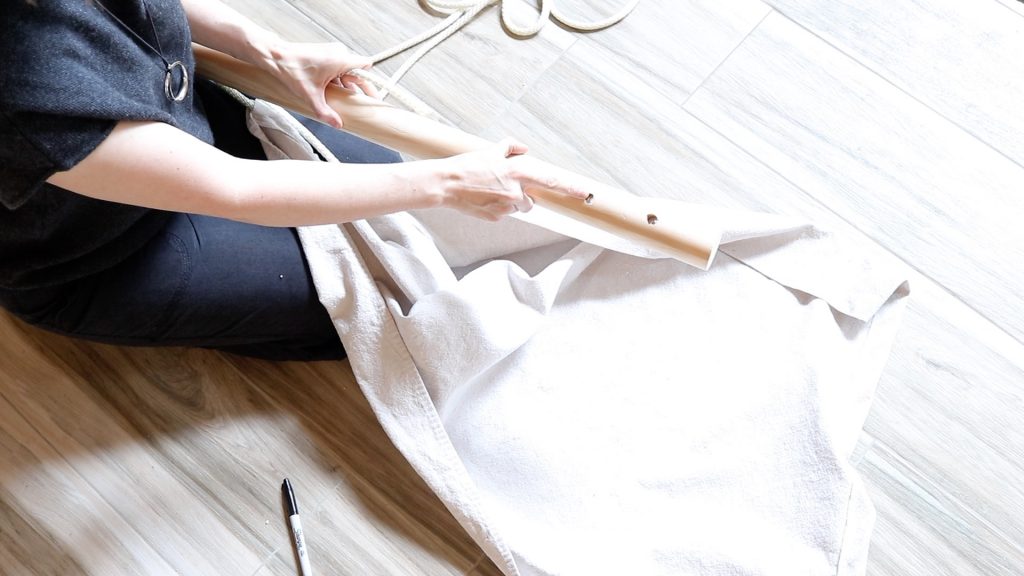 A woman is holding a dowel with holes drilled in it and pointing to the hoe that the rope needs to be fed through for the next step of constructing the hammock chair.