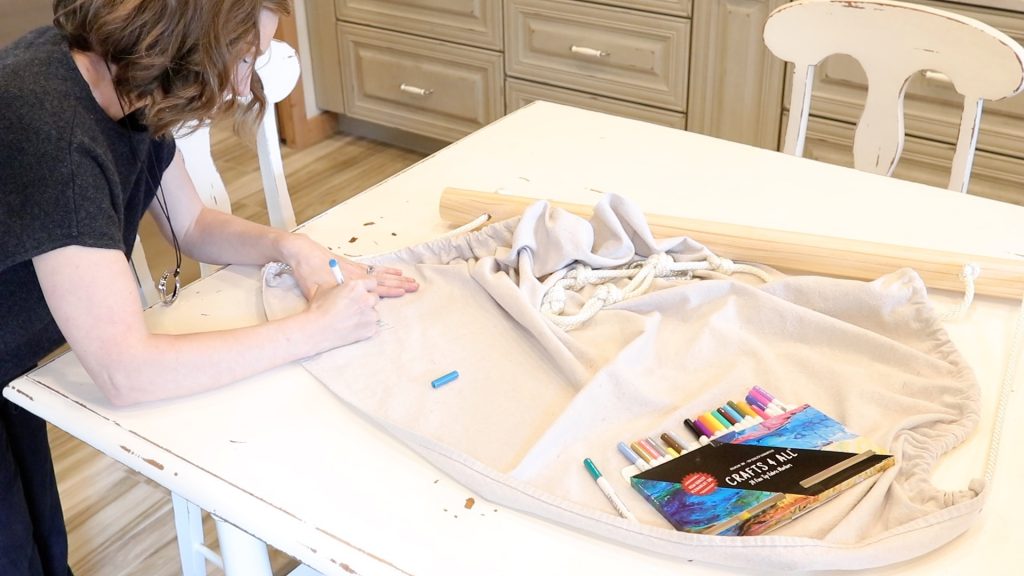 A woman is using a box of fabric pens to color on her DIY fabric swinging chair. The chair is laying on a white table.