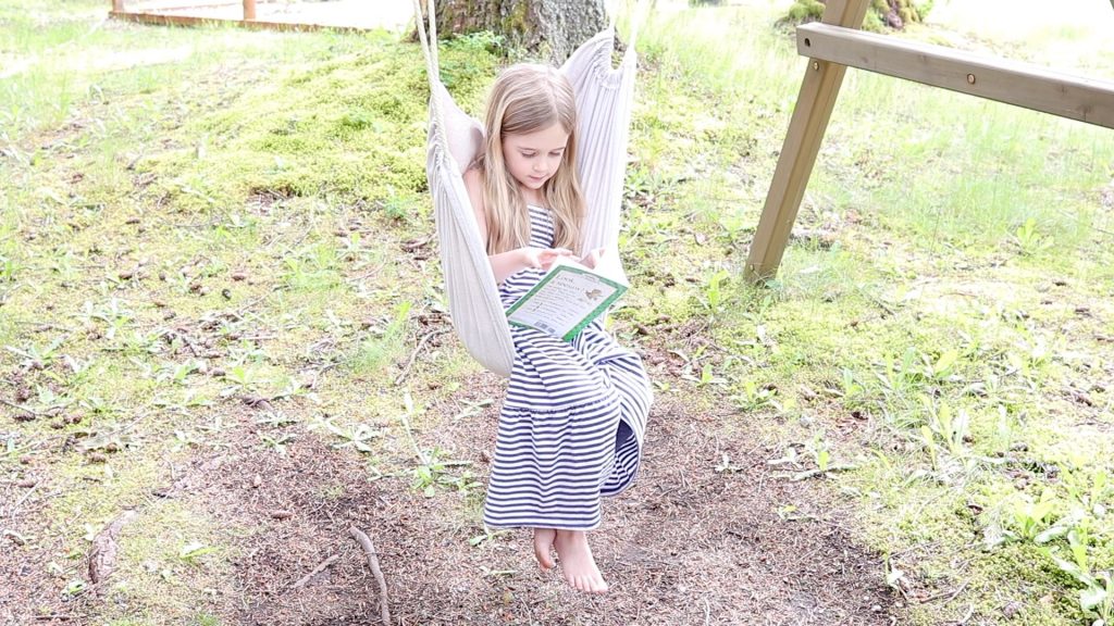 A little girl sitting in the DIY swing chair that her mom just finished making. She is reading a book and wearing a navy blue and white striped dress. The sky chair is hanging outside from an old swing set.