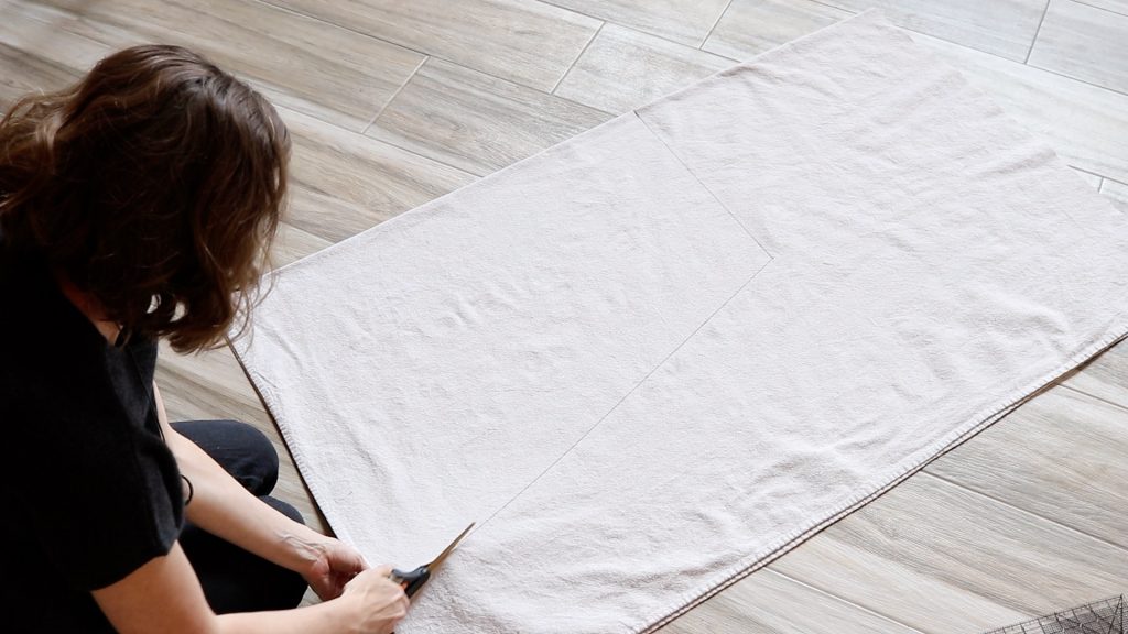 A woman with brown hair is leaning over the large piece of canvas fabric and cutting it to size with a pair of scissors. 