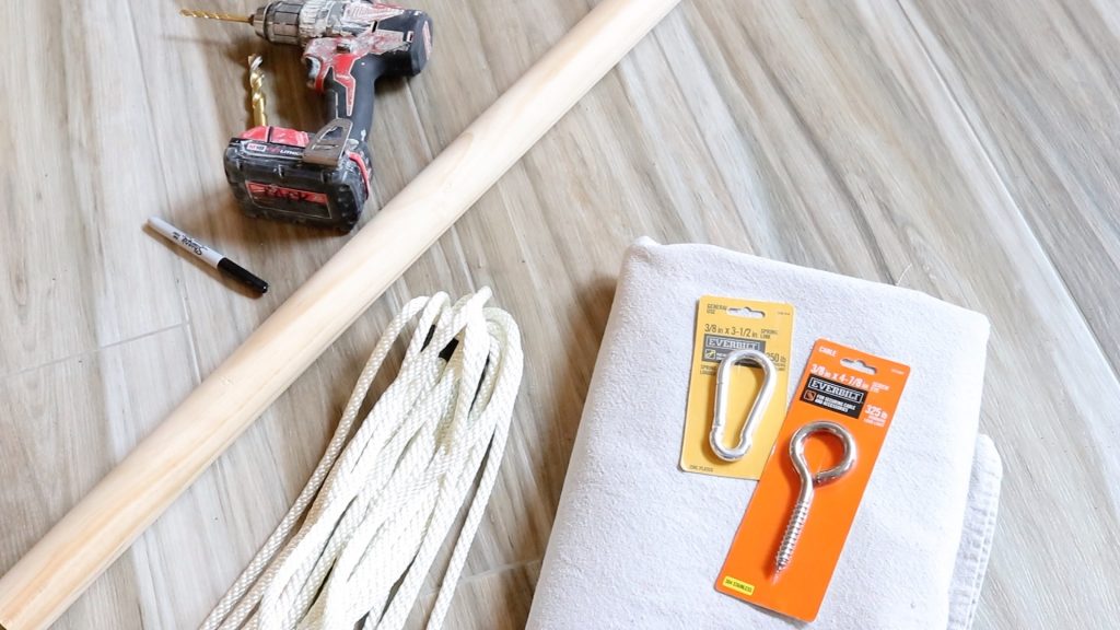 A picture of the supplies needed to make a DIY sky chair. A drill, sharpie marker, canvas fabric, an eye bolt and rope are laying on a wooden table. 