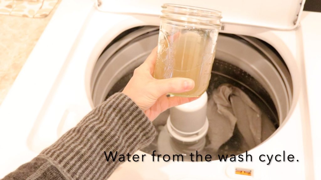 A womans hand holding a dirty glass of water from the canvas washing process. The purpose is to demonstrate the need to wash the fabric prior to use. 