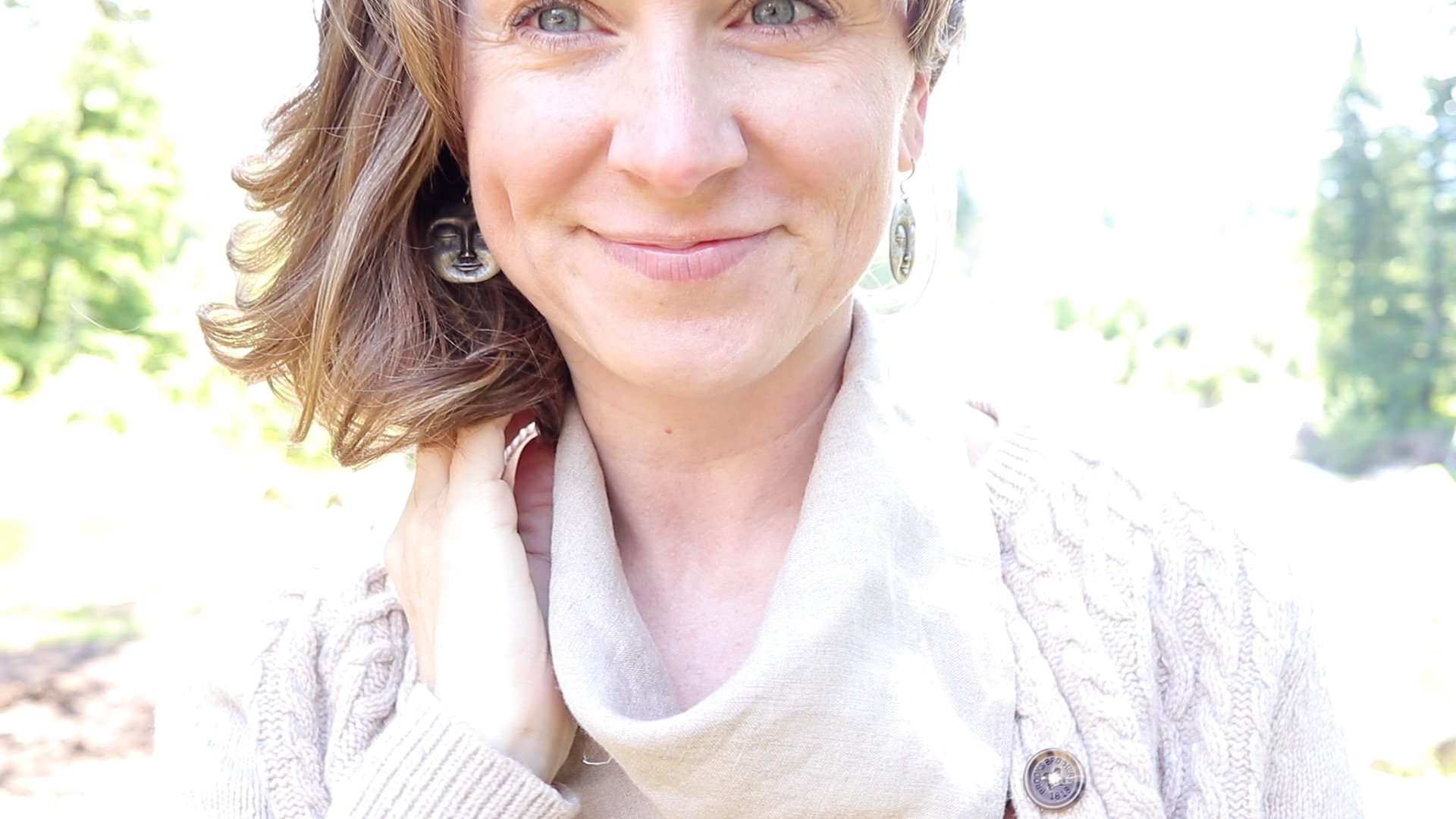 Girl smiling at the camera standing in the sunshine. She's wearing a tan well cable knit sweater and a tan linen bandana