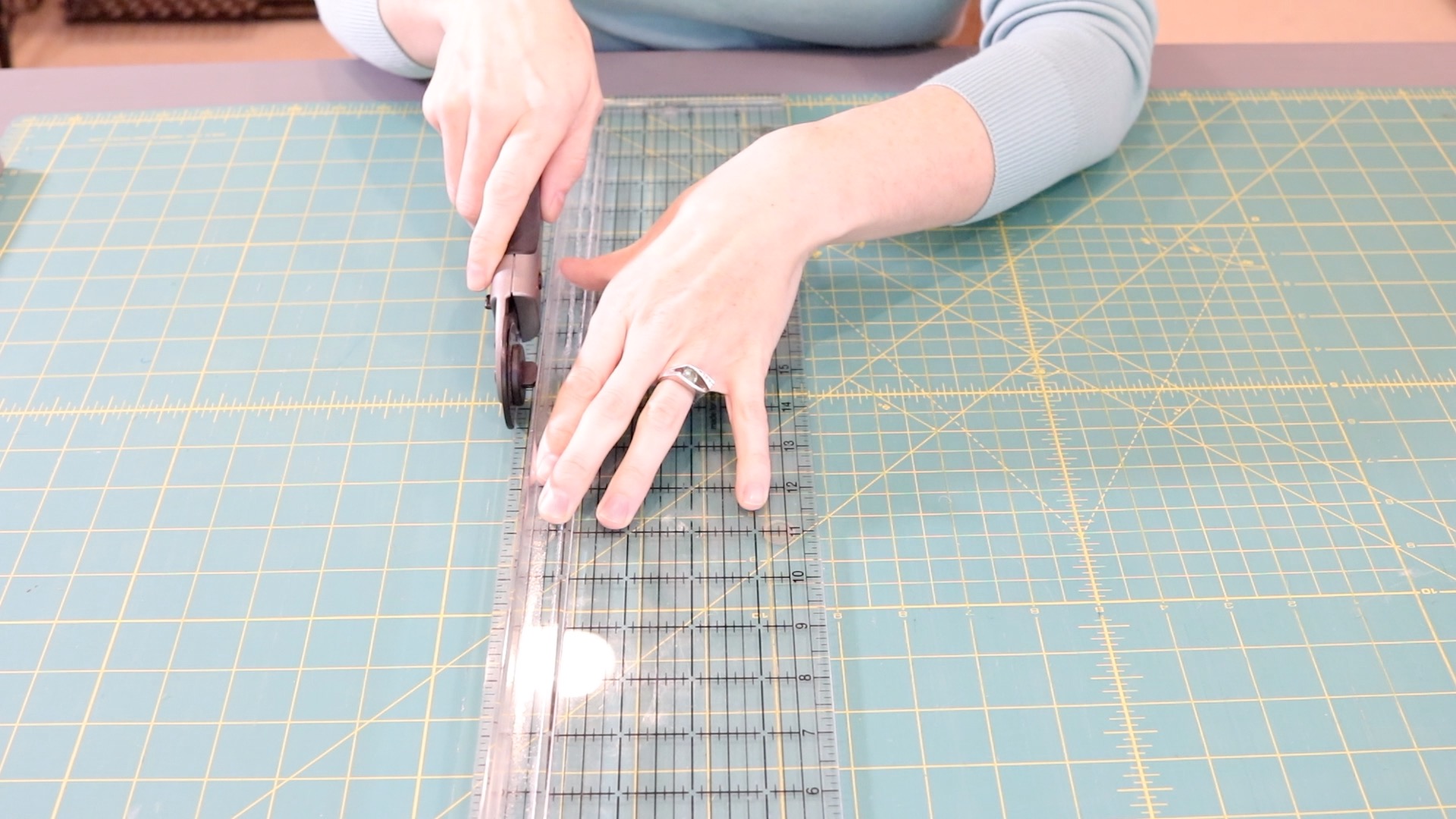 A girl wearing a blue shirt demonstrating how to hold the ruler and rotary cutter while cutting fabric on a cutting mat.