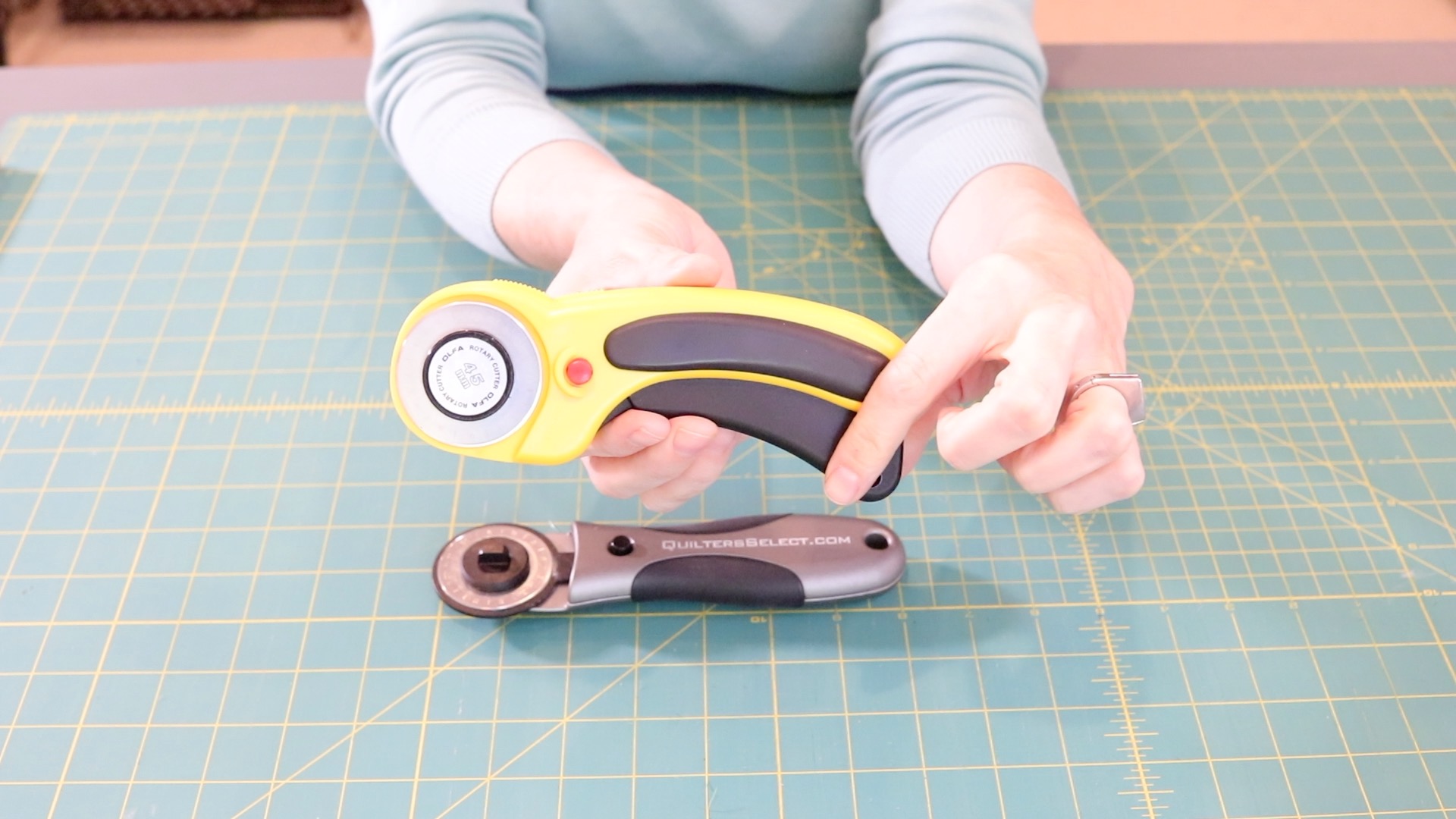 A hand holding a yellow rotary cutter while a gray metal cutter sits on a green cutting mat below. 