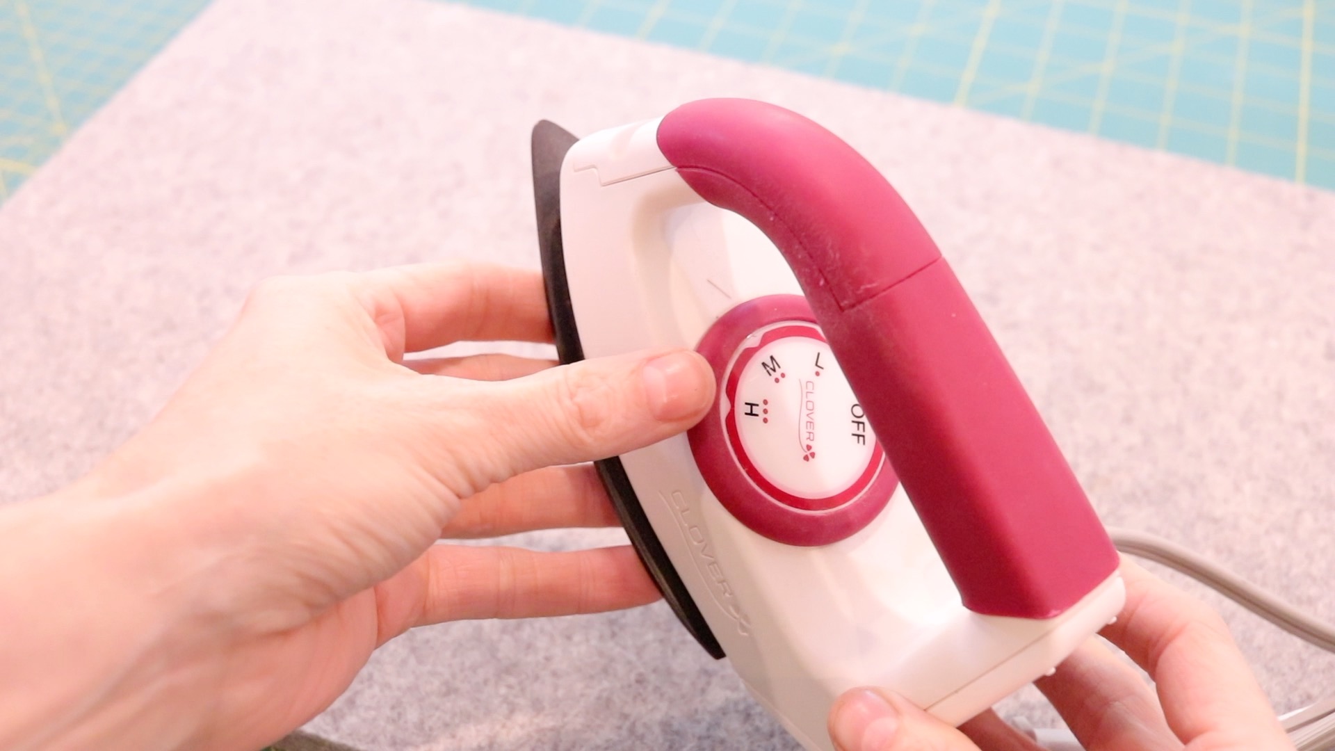 A close up of a mini clover sewing iron and its temperature setting features. The iron is the size of a hand and is fuchsia and white.