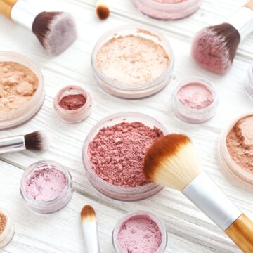 a white barn board tabletop with several containers of blush and eyeshadows in varying shades of soft pink hues. makeup brushes surround the delicate containers of powdered products