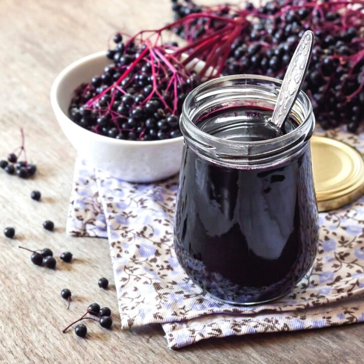 a jar of freshly made thickened elderberry syrup. The clear jar of syrup sits on a table top surrounded by fresh picked elderberries . A silver spoon sits in the jar to stir.