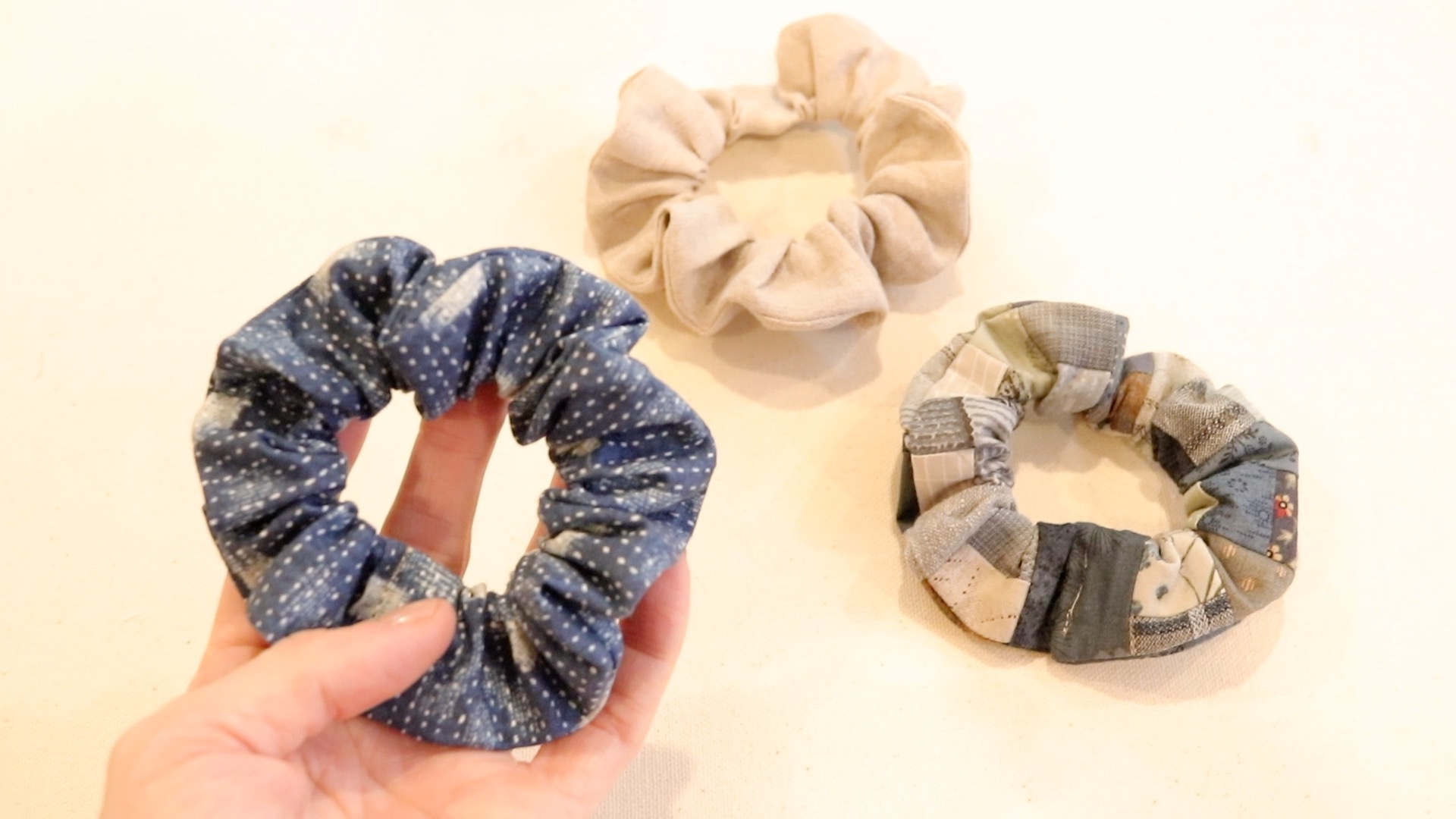 Three homemade hair scrunchies resting on a table. A womans hand is holding a blue and white speckled scrunchie up towards the camera to see the pattern on the fabric.