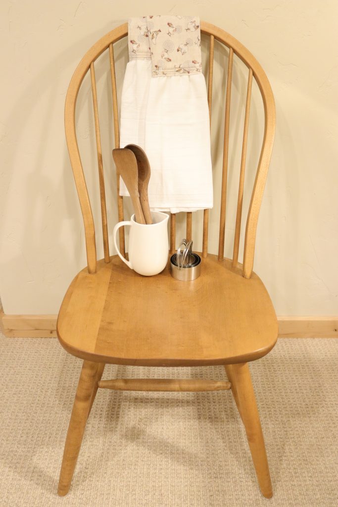 A newly completed homemade kitchen scarf laying over the back of a wooden chair. A white cup with wooden spoons and biscuit cutters rest also on the chair. The chair is resting next to a cream colored wall.