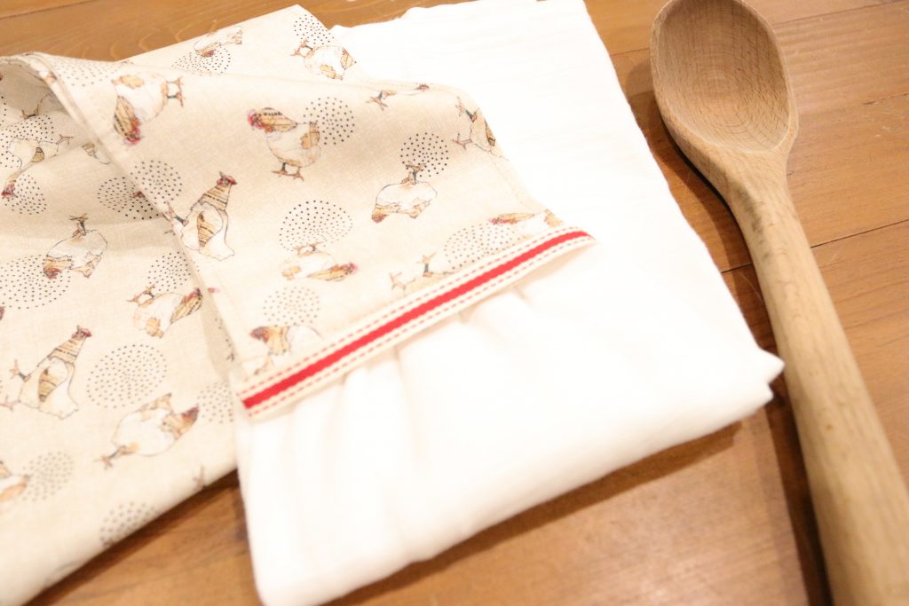 A homemade kitchen scarf sitting on a wooden table next to a wooden spoon. The towel is Mae from a chicken printed fabric with white gauze fabric at the bottom.