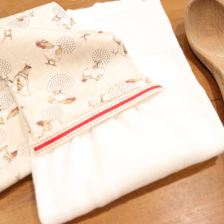 a kitchen scarf sewn from a chicken printed fabric with a white tea towel. A red and white piece of ribbon divides the two pieces of fabric. The kitchen boa is sitting on an antique table with a wooden spoon sitting near by