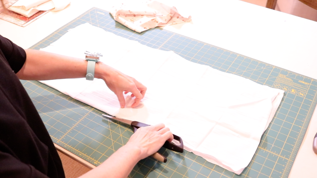 A woman finishes preparing the white gauze fabric for her sewing project by cutting it in half with a pair of black handled scissors. 