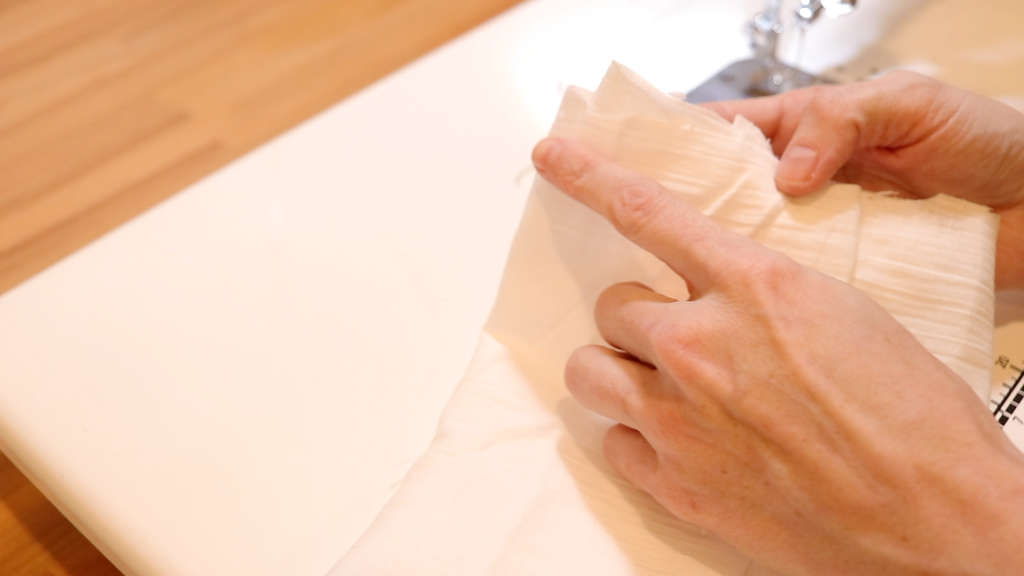 Folding and pleating white gauze fabric for the kitchen scarf. A womans hand is pointing to where the seam on the fabric will be sewn after pleating. 